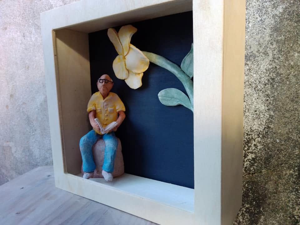 Small seated clay figure of a man in a yellow shirt inside a wooden box, with a large yellow flower in relief behind him