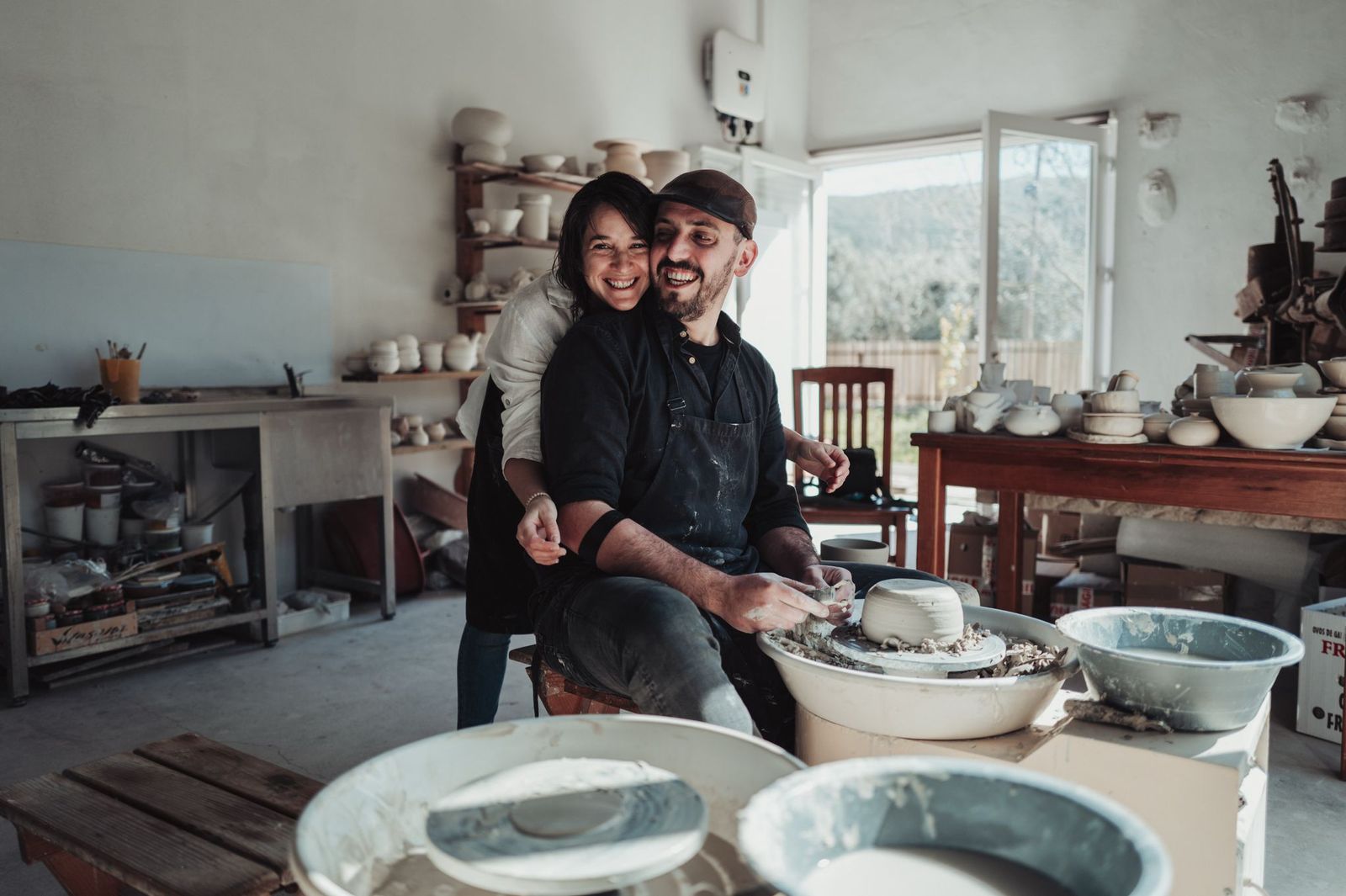 José and Diana Siphioni laughing together at the potter's wheel in their studio, shelves of pieces behind them
