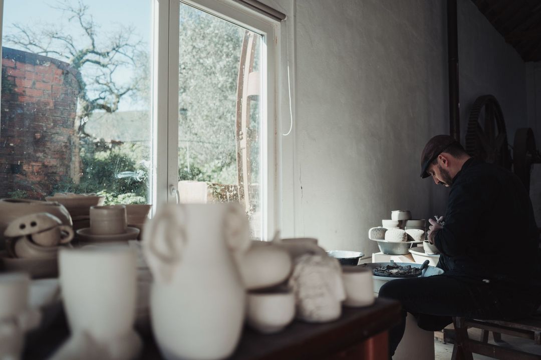 José working by the window, unfired jugs and cups on the shelf in front