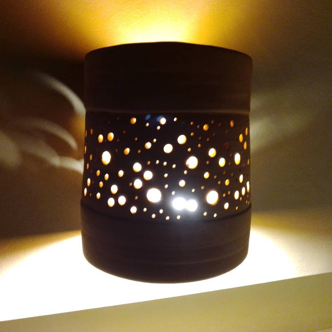 Dark cylindrical ceramic lamp pierced with small holes, the light shining through