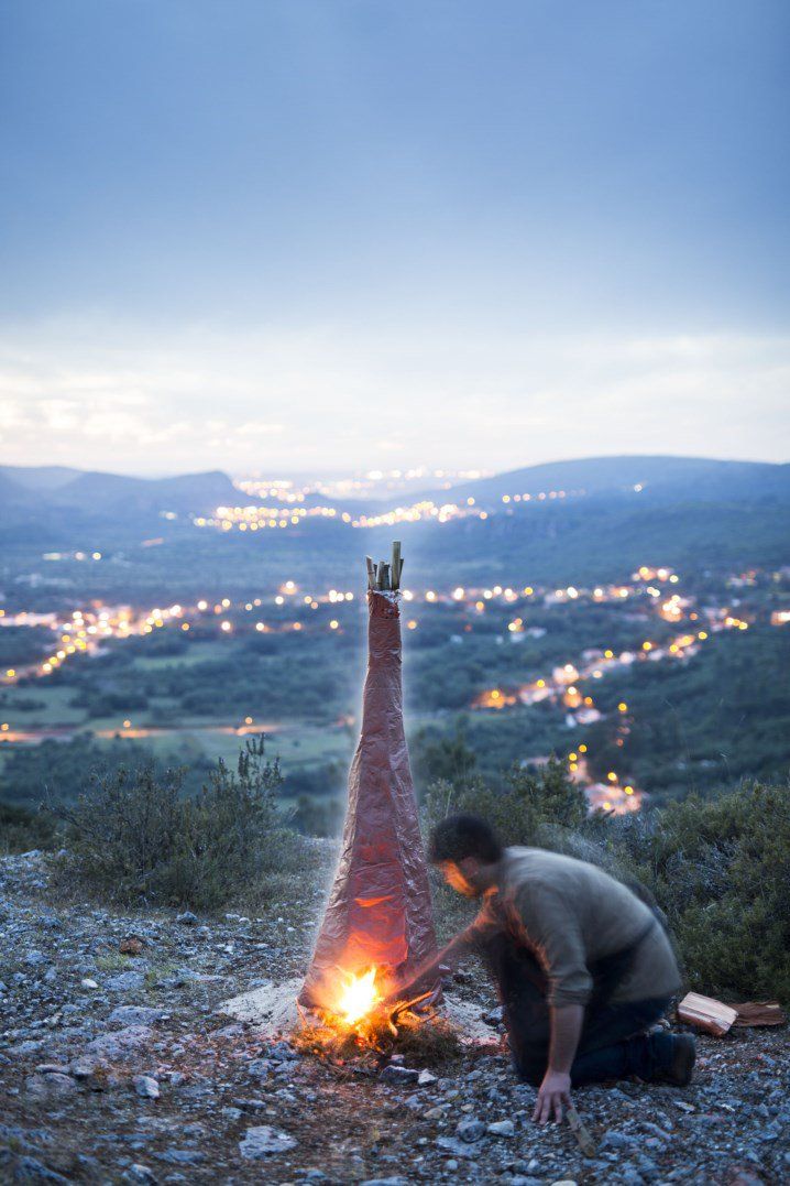 A potter kneeling to light a paper kiln on a hillside at dusk, the lights of the valley behind him