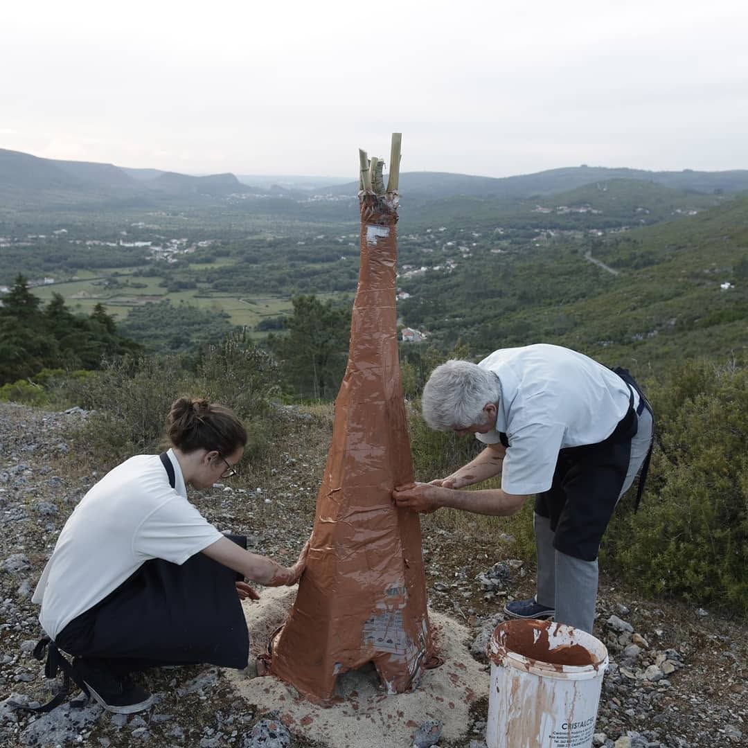 Dois participantes a construir com barro um forno de papel alto numa encosta sobre o vale
