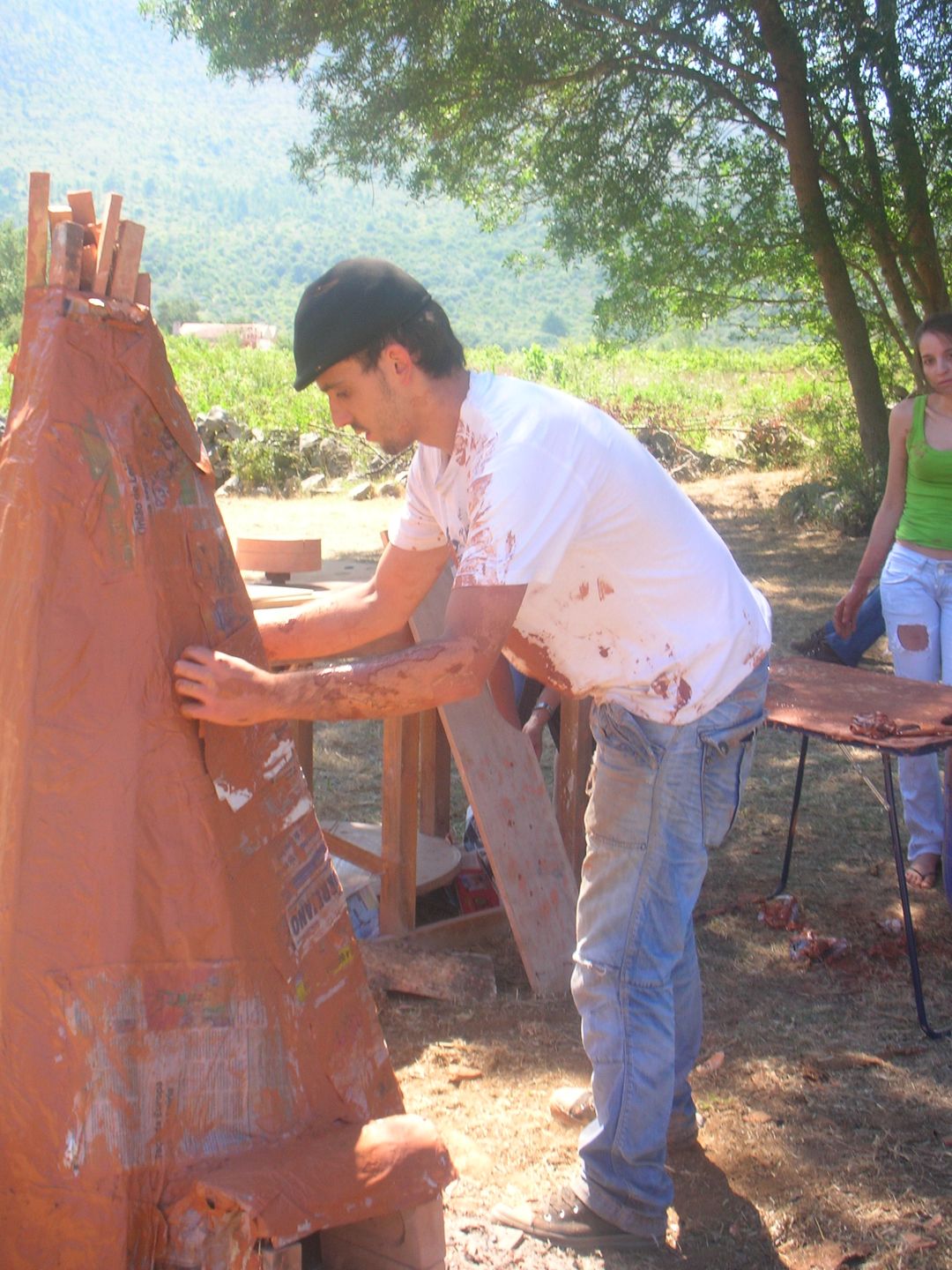 A man covering a tall, cone-shaped paper kiln with wet clay outdoors