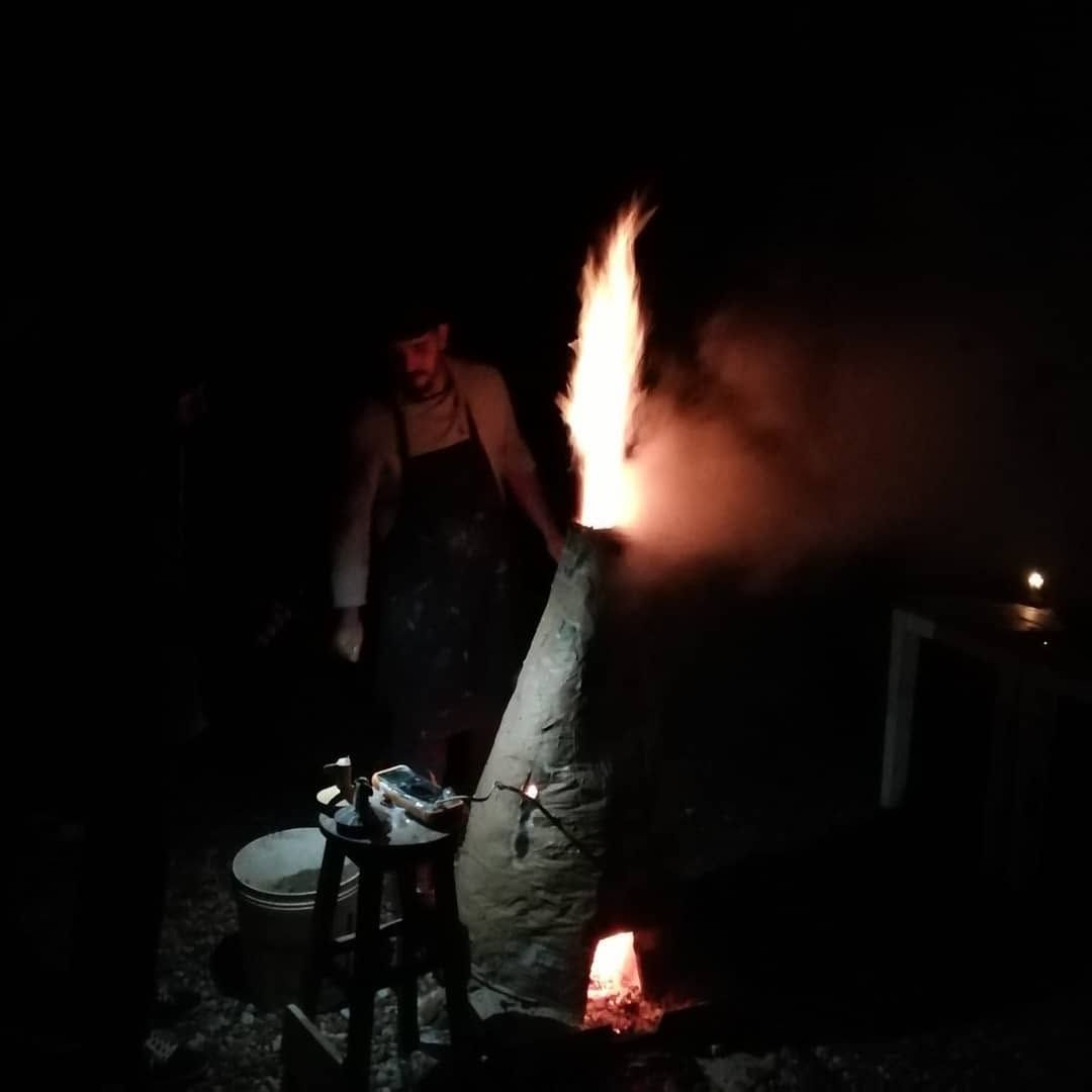 A tall paper kiln burning in the dark, a flame rising from the top while a potter watches the fire