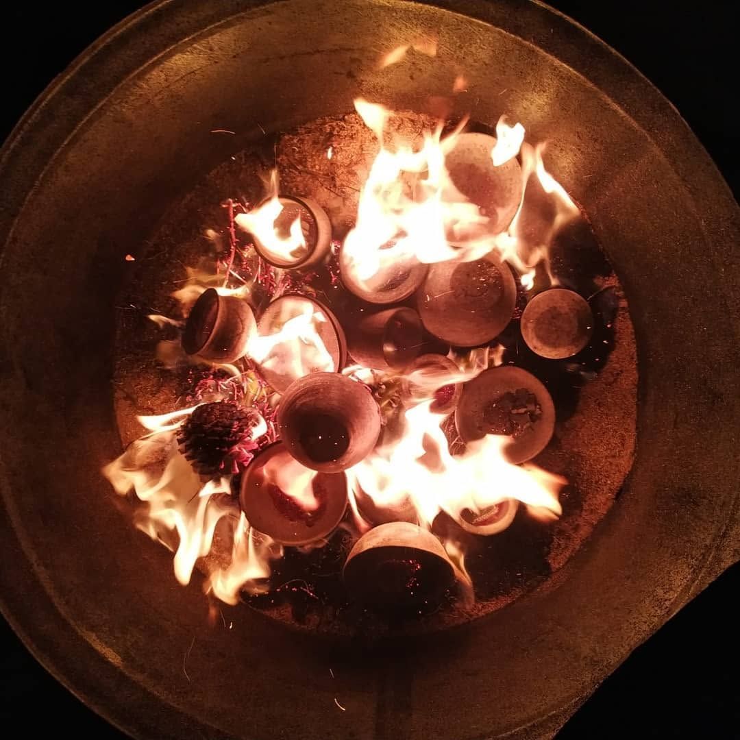 Raku firing at night: small clay cups and bowls among flames and pine cones in a metal barrel, seen from above