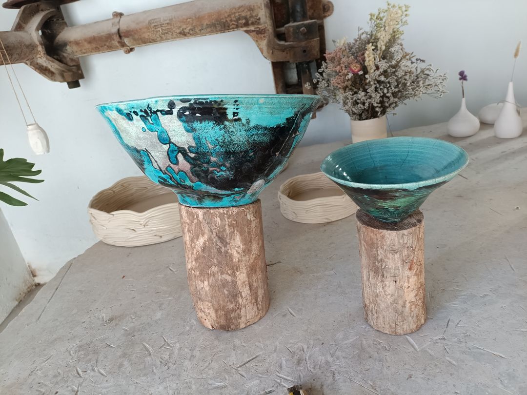 Two wide bowls with a turquoise and black glaze, each standing on a short log, dried flowers behind